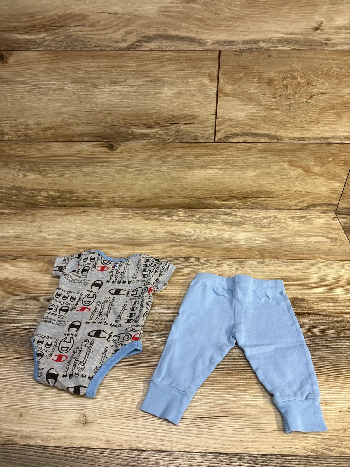 Flat lay of Champion gray printed short-sleeve bodysuit and light blue pants, size 3M, showing front view, snap closures, and elastic waistband on wood background