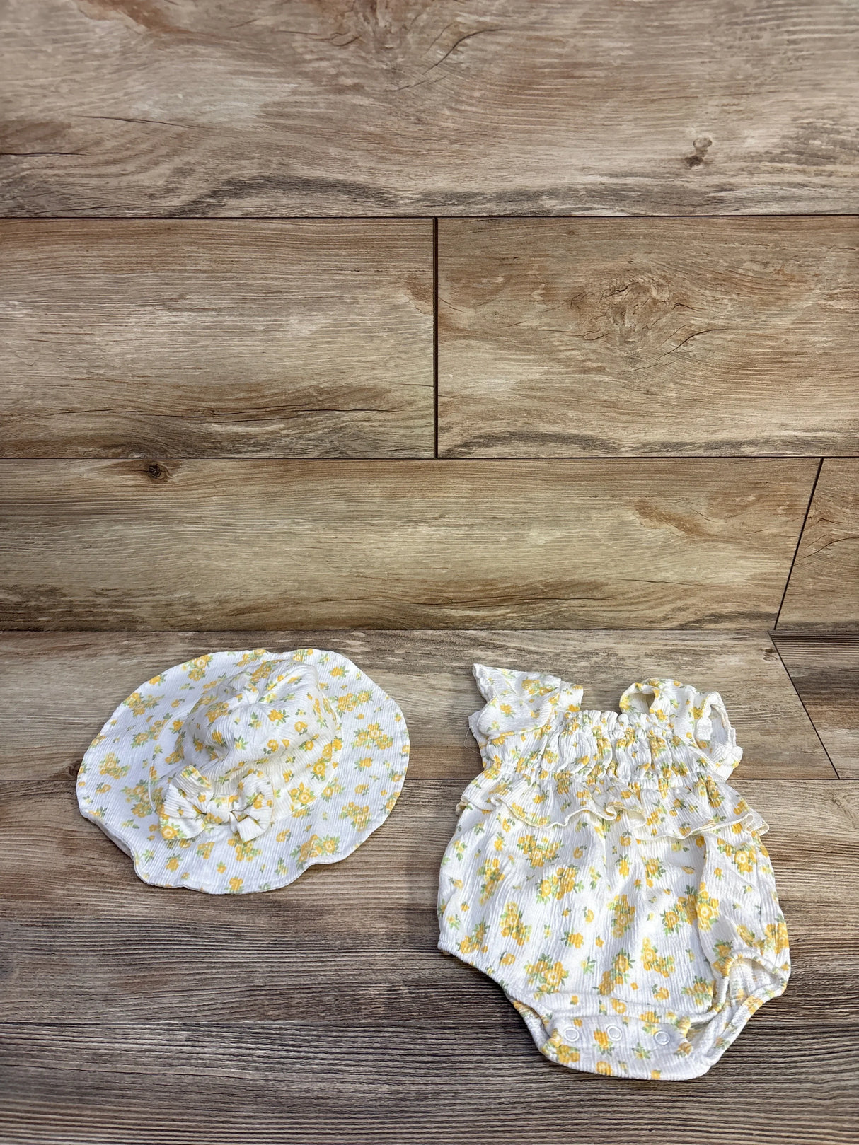 Top-down view of white 0-3M floral smocked bubble romper and matching sun hat showcasing ruffled smocking, snap crotch and yellow flower print on wood background