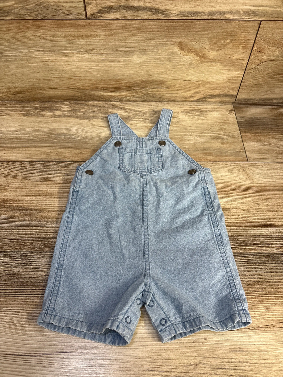 Front view of Carter’s blue chambray shortalls showcasing adjustable straps, front pocket, and snap closures on wood flooring