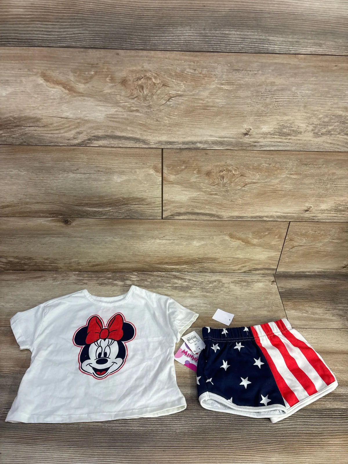 Flat lay of toddler white Minnie Mouse tee showcasing front Minnie graphic with navy and red stars and stripes shorts on wood floor