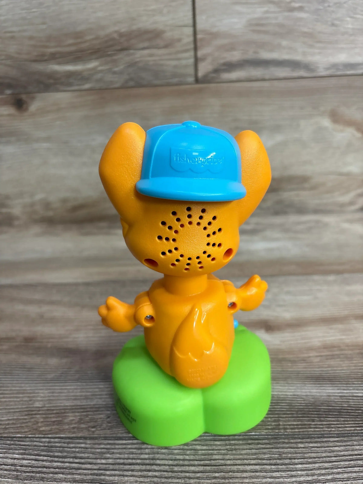 Back of a yellow Fisher-Price toy character with blue hat on green base, wood background