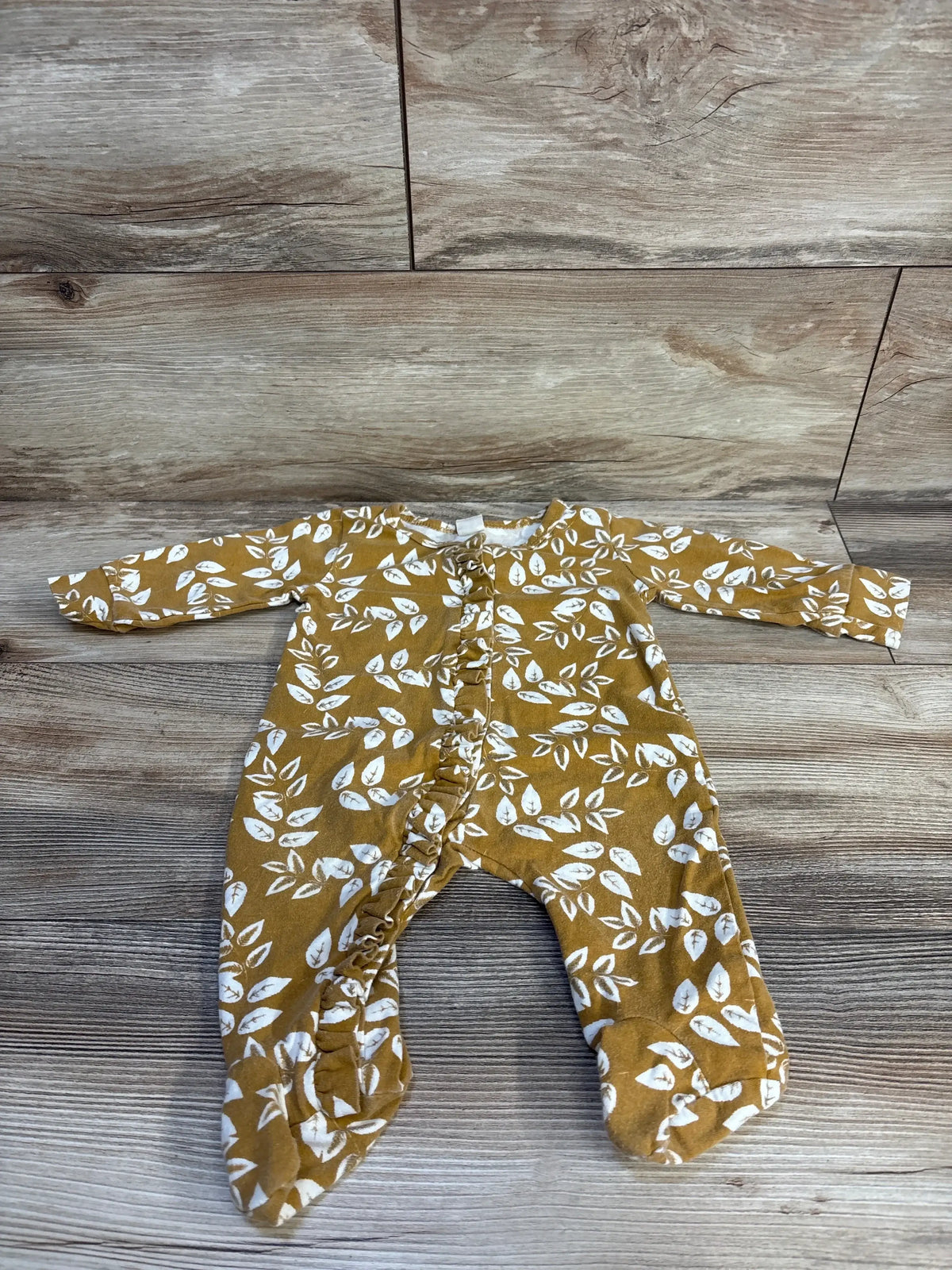 Mustard baby onesie with white leaf print, ruffled legs, laid on wood floor