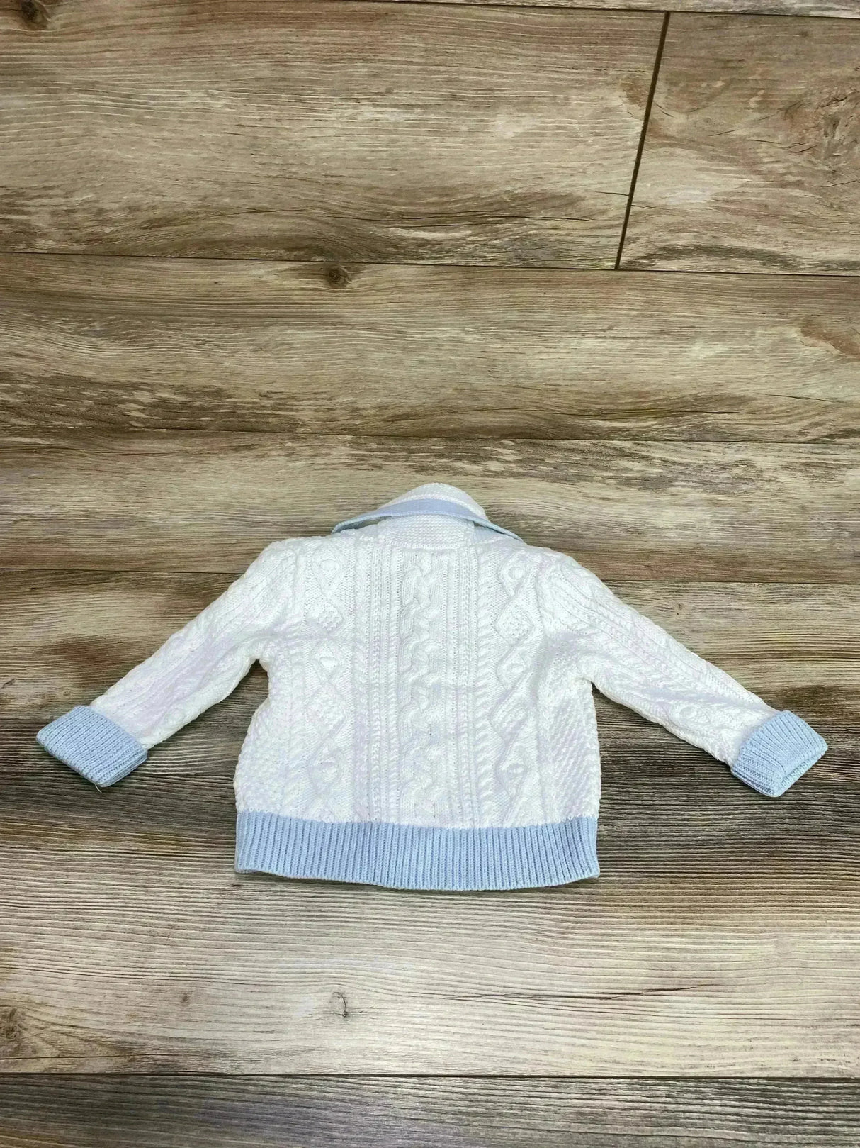 Back view of baby shawl neck cable knit cardigan with cable pattern and light blue cuffs and hem on wood background