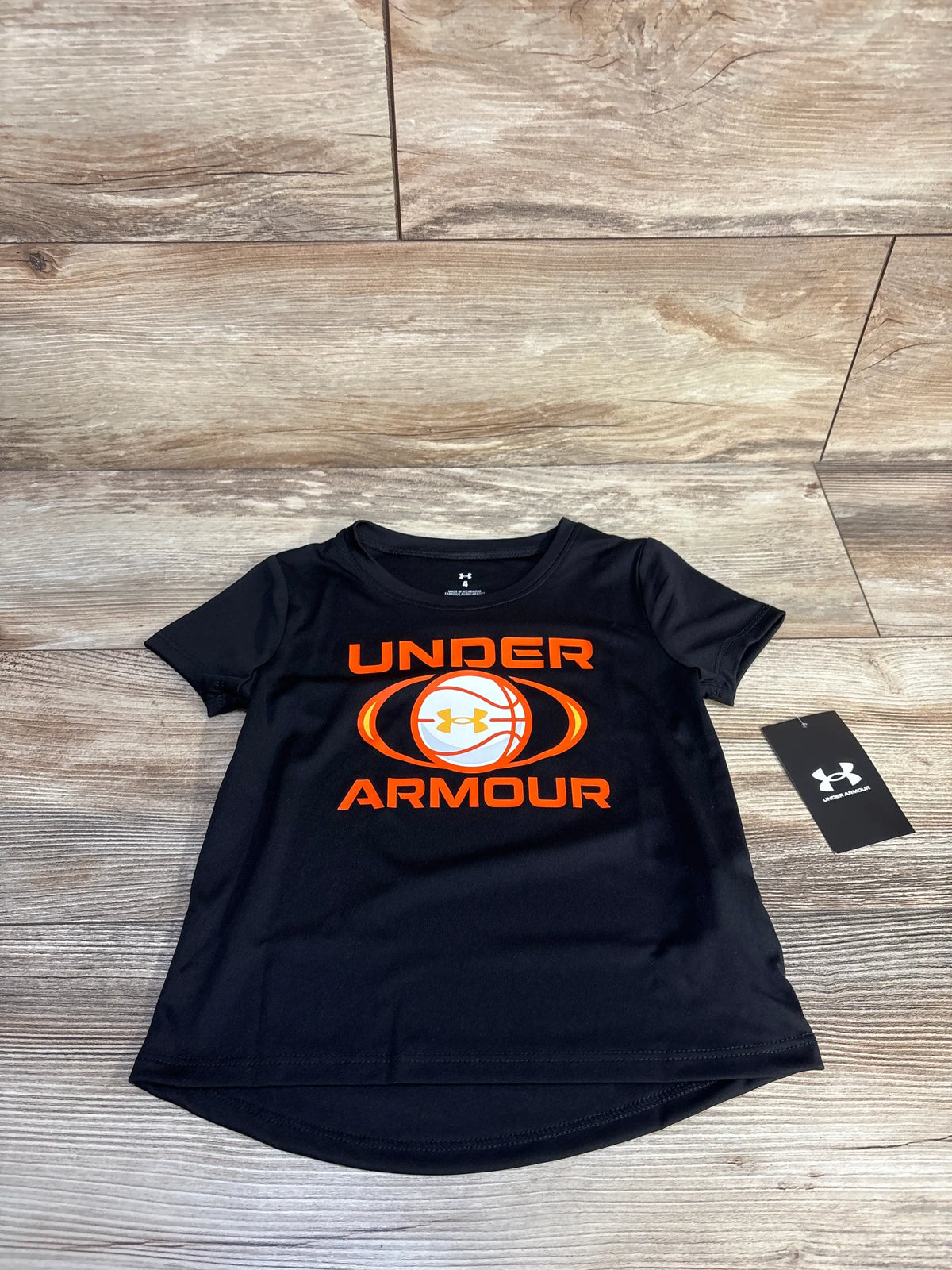 Front view of black Under Armour Velocity Ball T-shirt showcasing orange basketball graphic on wood-patterned background