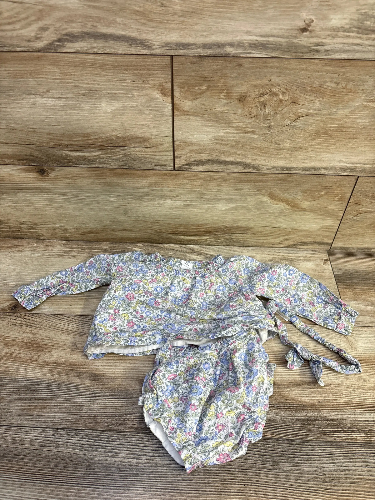 Top-down view of a white floral three-piece baby outfit with long-sleeve top, bloomers, and headband on a wood floor