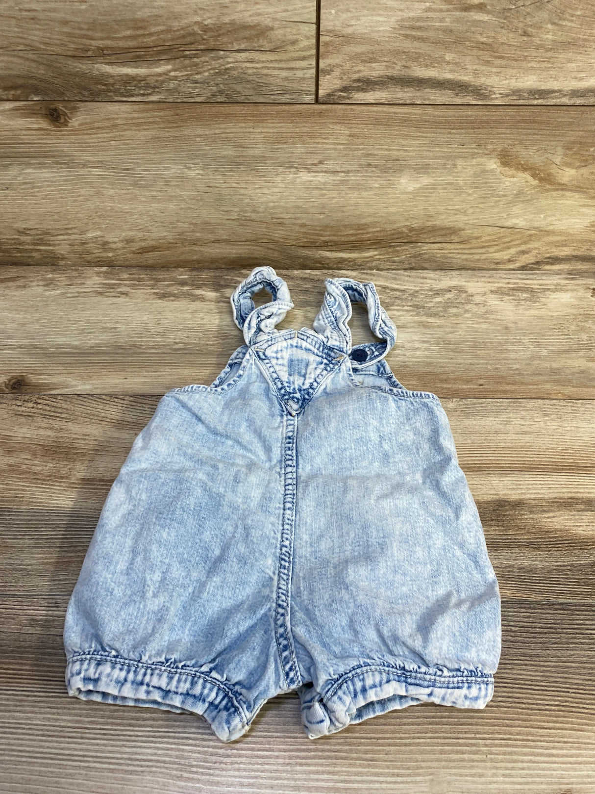 Front view of Baby Gap light blue denim shortalls with ruffled straps, faded wash and elasticized hems on wood background