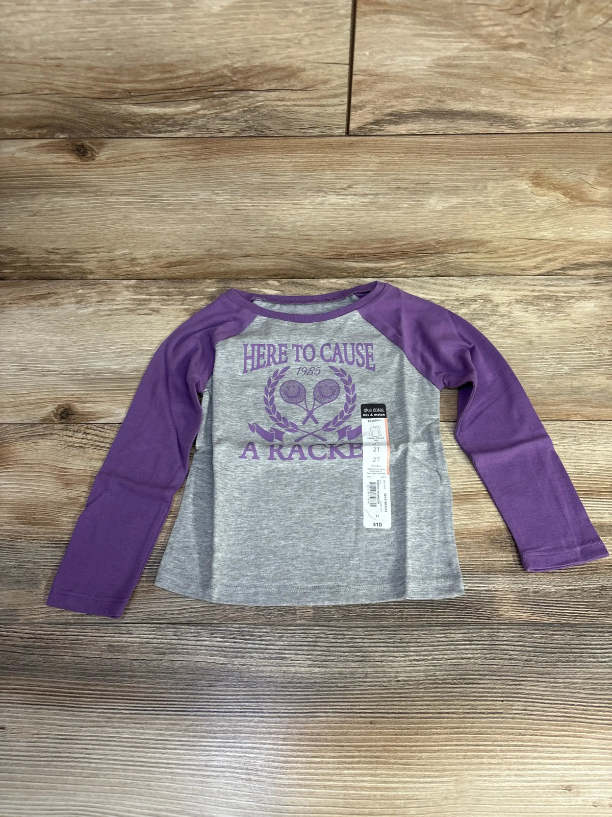 Flat lay of grey raglan toddler tee with purple sleeves Here To Cause A Racket tennis graphic and 2T hangtag on wood floor