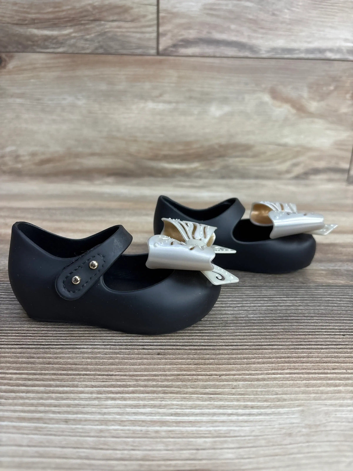 Side view of Mini Melissa Ultragirl Bow jelly flats, black size 5C, showing pearl bows and Mary Jane strap on wood