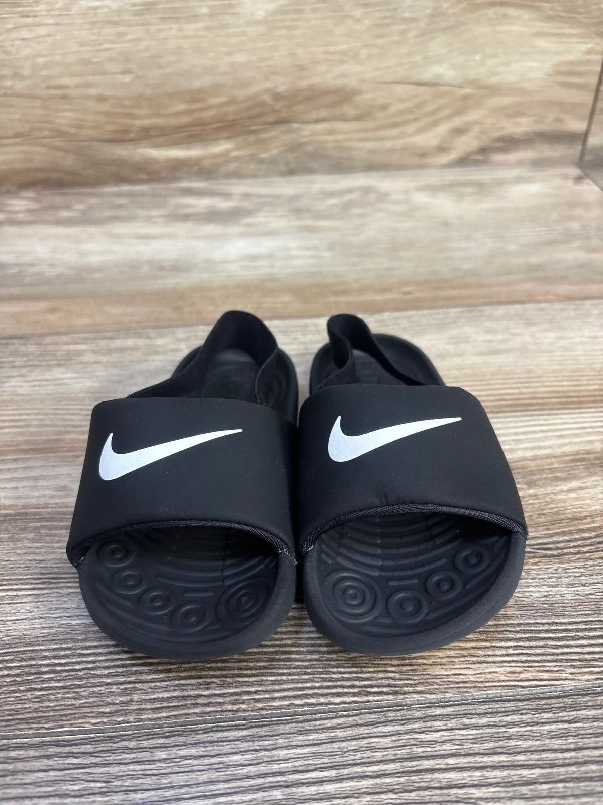 Front view of size 7C Nike Kawa black toddler slide sandals showcasing white swoosh on strap, textured footbed, and molded heel on wood floor