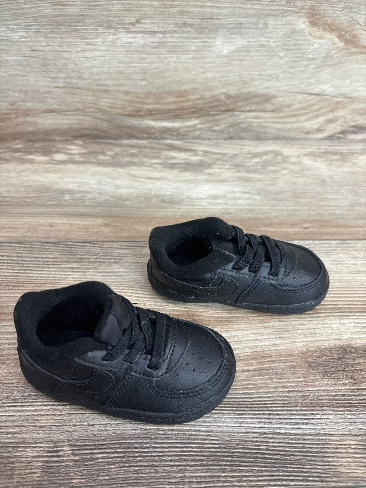 Angled top view of baby Nike Air Force 1 crib shoes showing black leather upper, laces, and padded collar on wood background