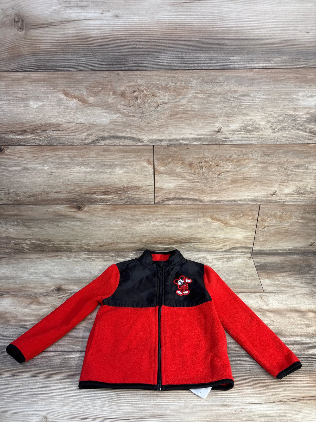 Front view of red toddler fleece jacket with black yoke and zipper, embroidered Mickey Mouse patch, size 4T, wood background