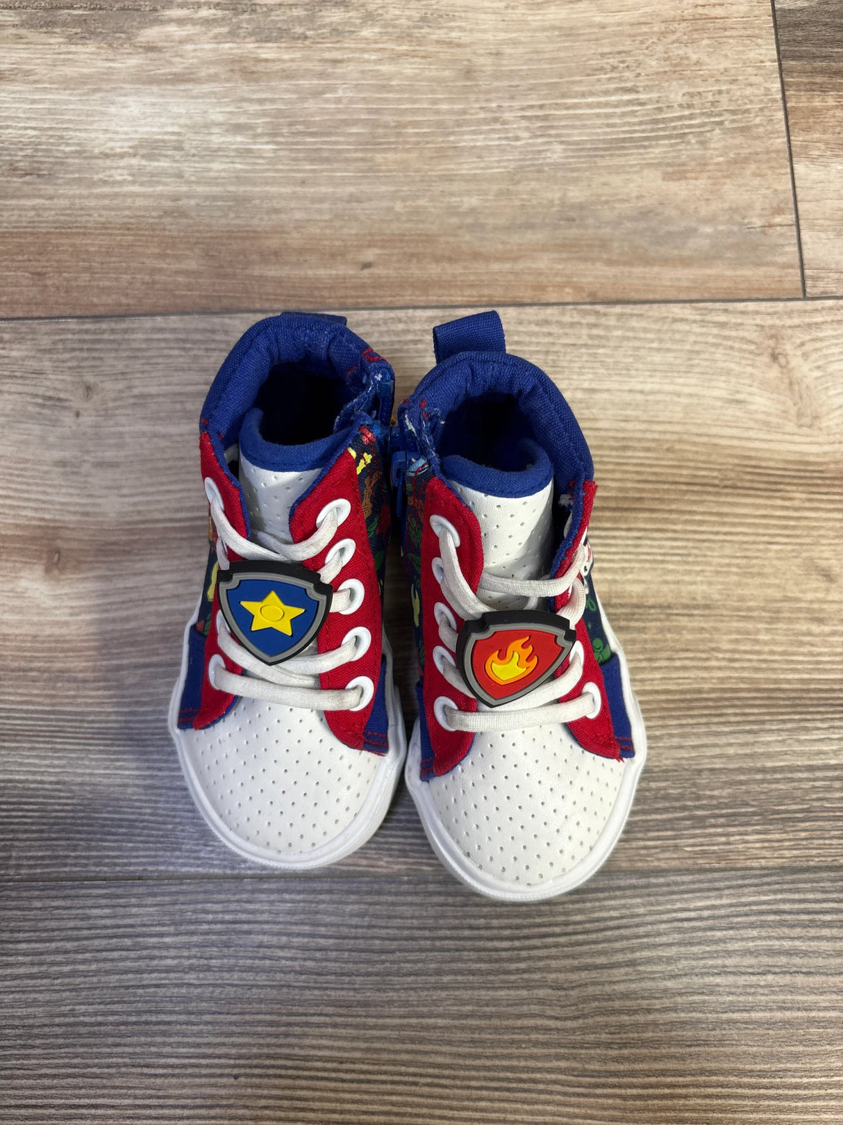 Top view of Ground Up Paw Patrol high top sneakers in white with red and blue panels and badge charms on wood floor