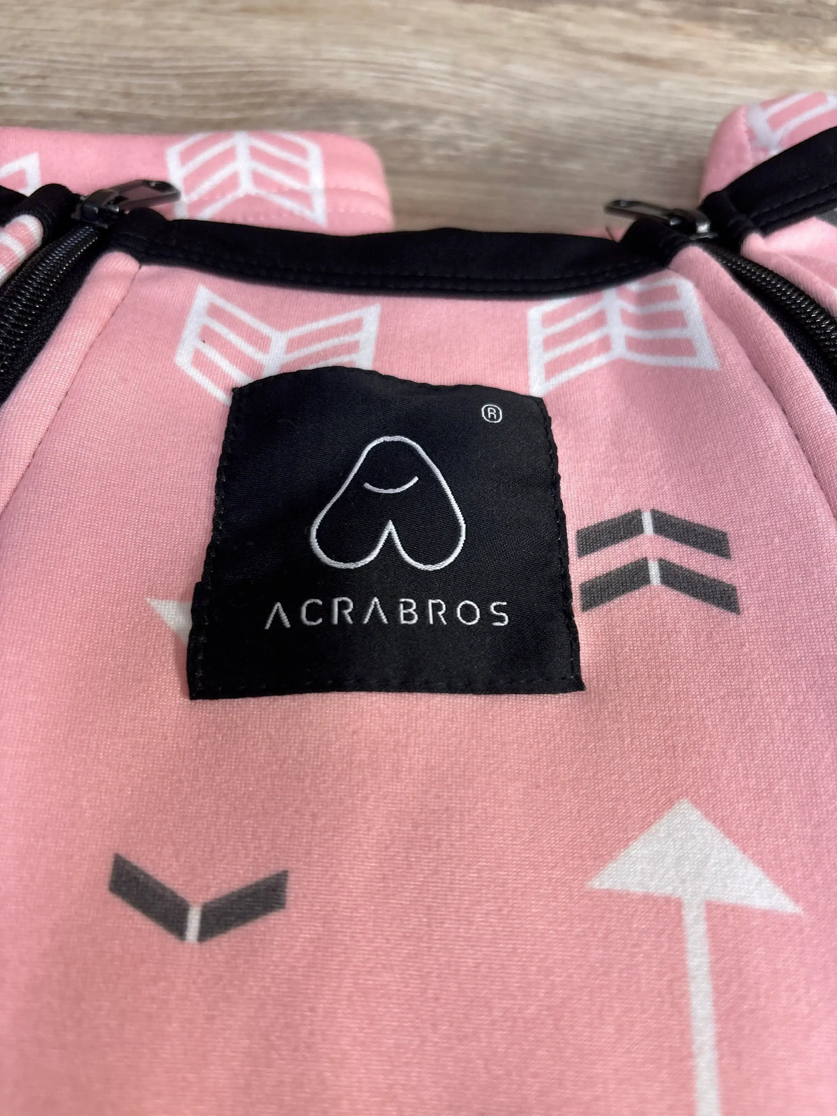 Close-up of Acrabros car seat cover showing black logo patch and white arrow pattern on pink fabric with wood background