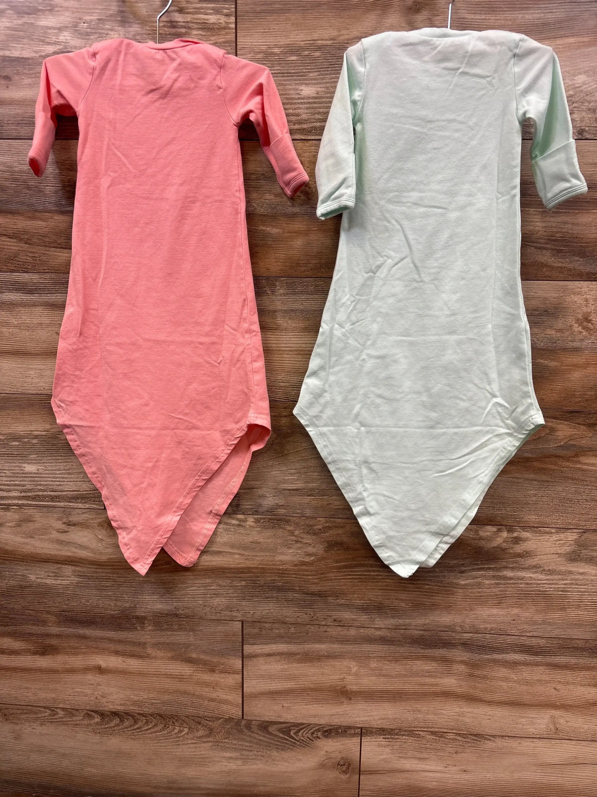 Hanging view of two Baby Noah knotted gowns size 3M in pink and light green showing long sleeves and knotted bottoms on wood background