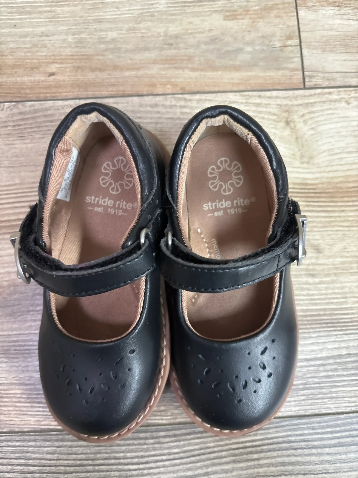Top view of Stride Rite Mara Mary Jane flats showcasing perforated toes and adjustable straps with a wood-look floor
