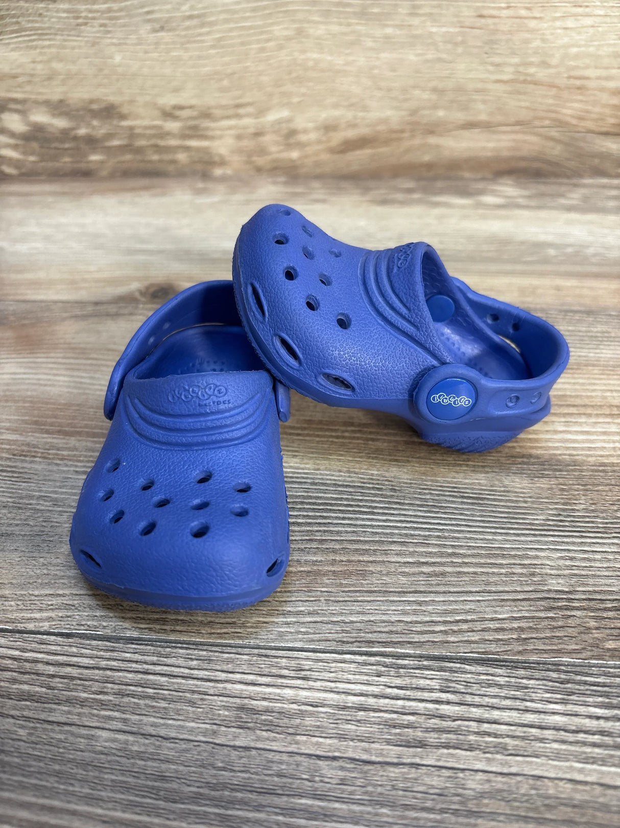 Three-quarter view of Crocs Classic Clog size 4/5C in blue showing ventilation holes, heel strap and textured footbed on wood