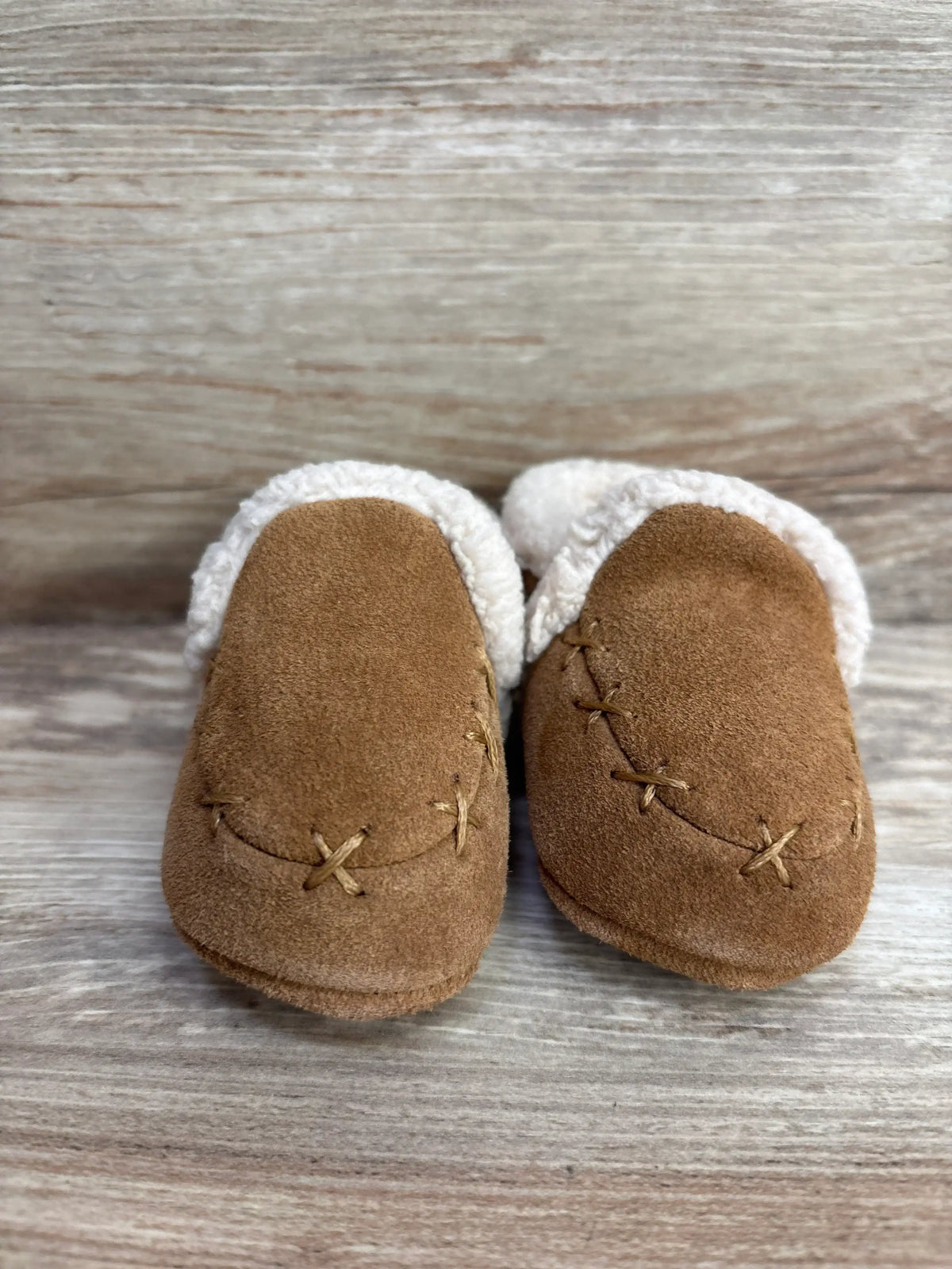 Brown suede baby moccasins with white fleece lining, displayed on wood background