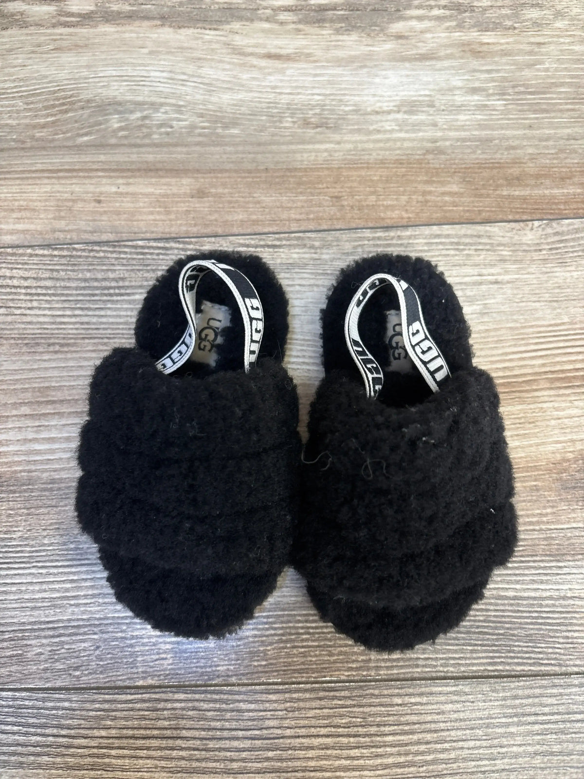 Top-down view of UGG Fluff Yeah Slides baby size 7C showing black fuzzy upper and UGG-branded elastic heel straps on wood background