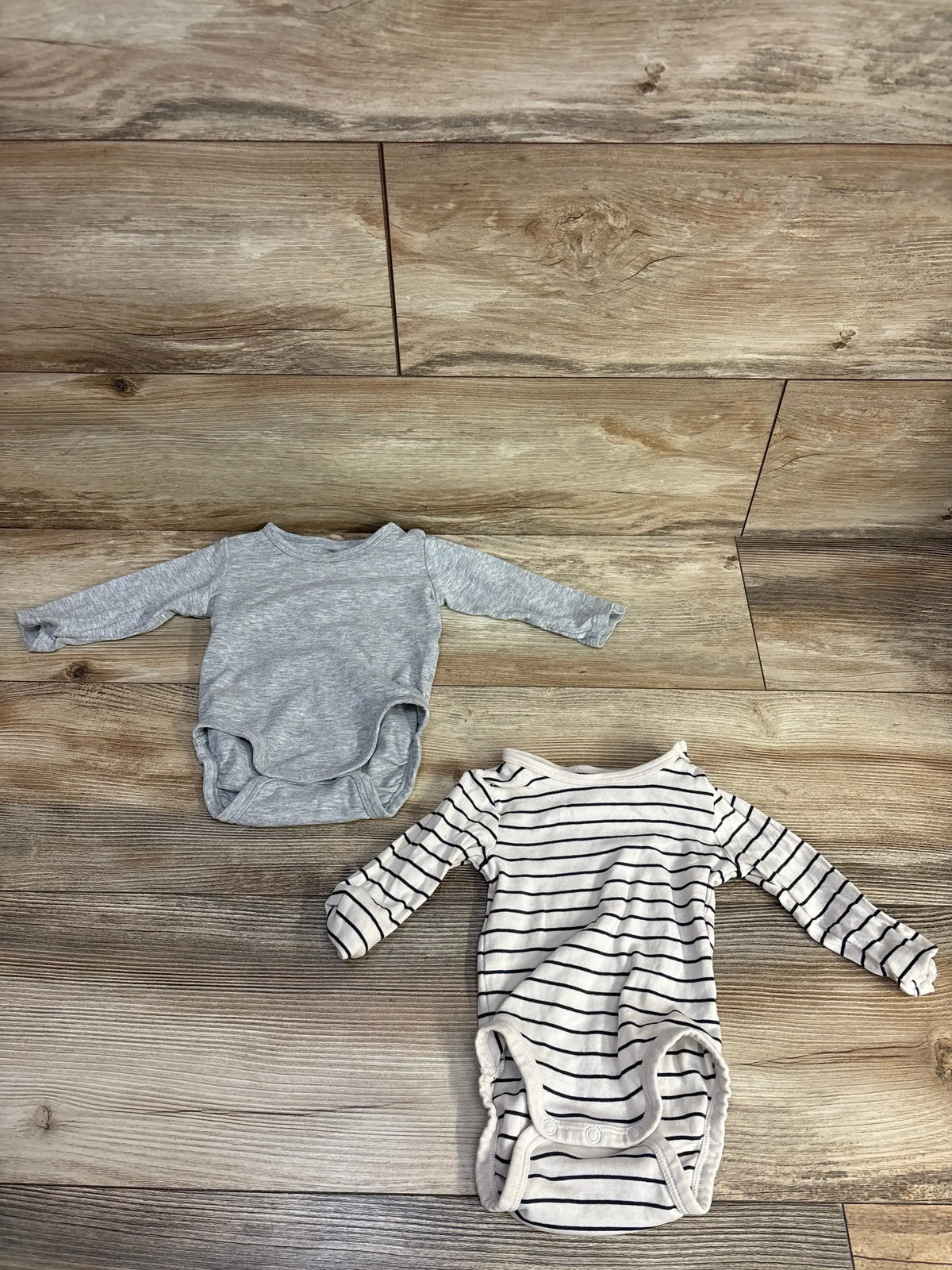 Two baby long sleeve onesies, one gray and one white with black stripes, on wooden floor