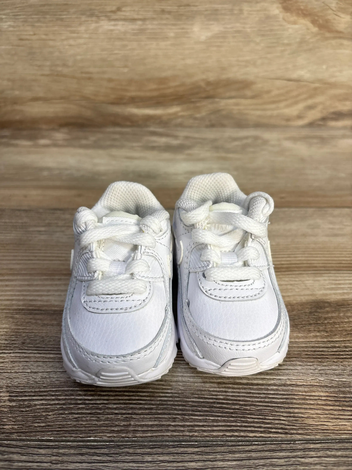 Front view of Nike Air Max 90 TD Triple White sneakers showing white laces and layered panels on wood grain