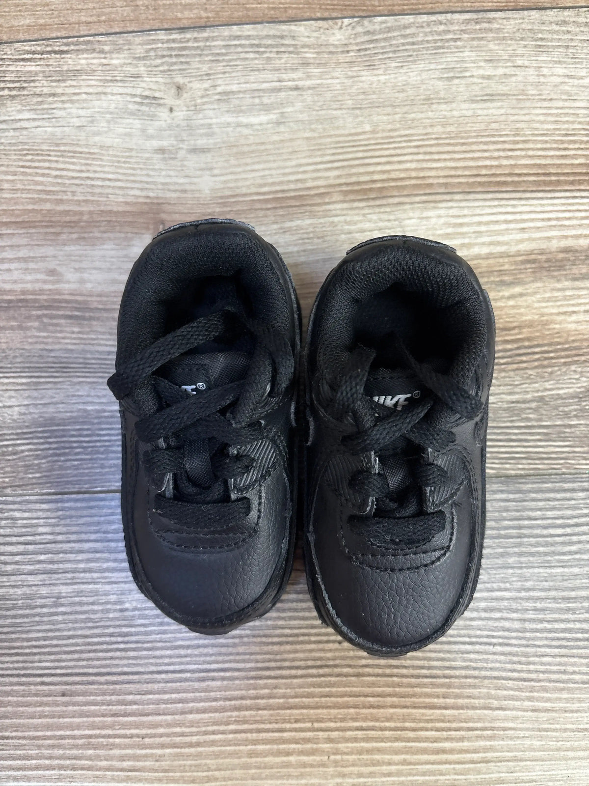 Top view of Nike Air Max 90 TD Triple Black toddler sneakers, size 4, showcasing black laces and padded collars on wood grain