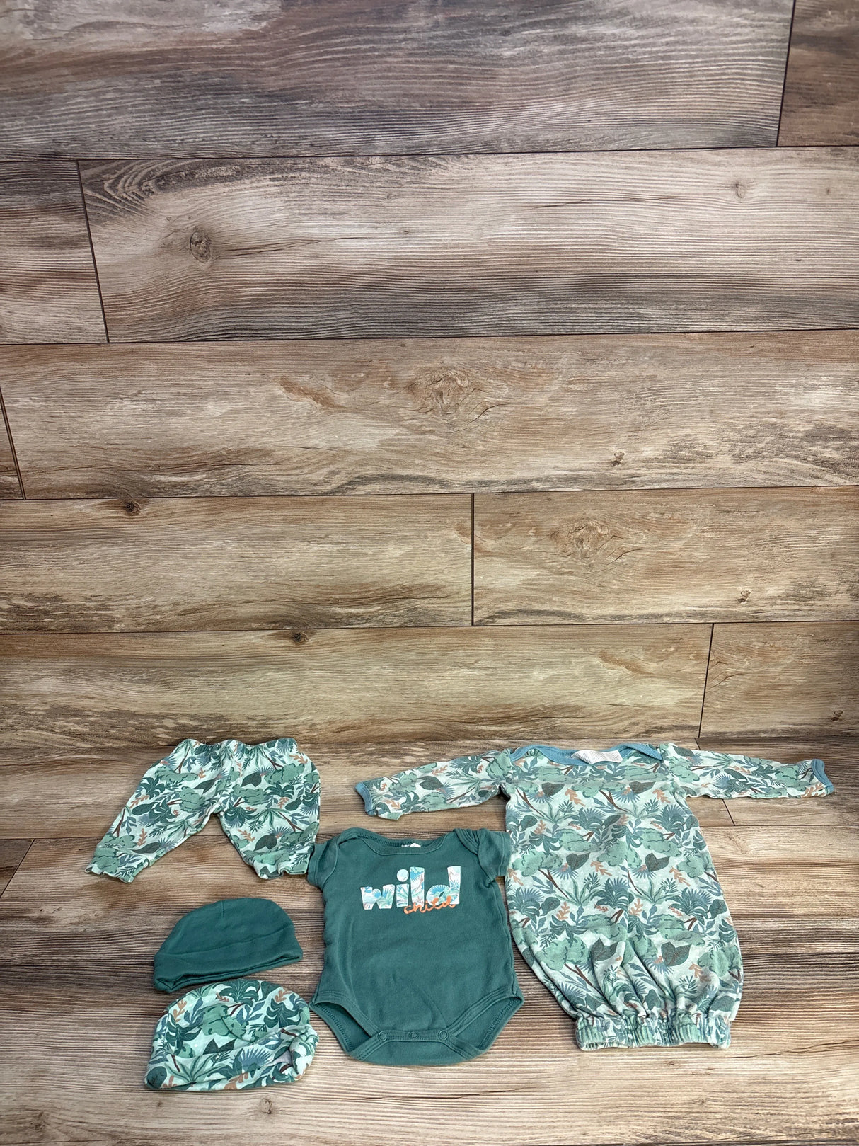 Baby outfit set with tropical leaf print, green onesie with 'wild' text, and matching hats on wood floor