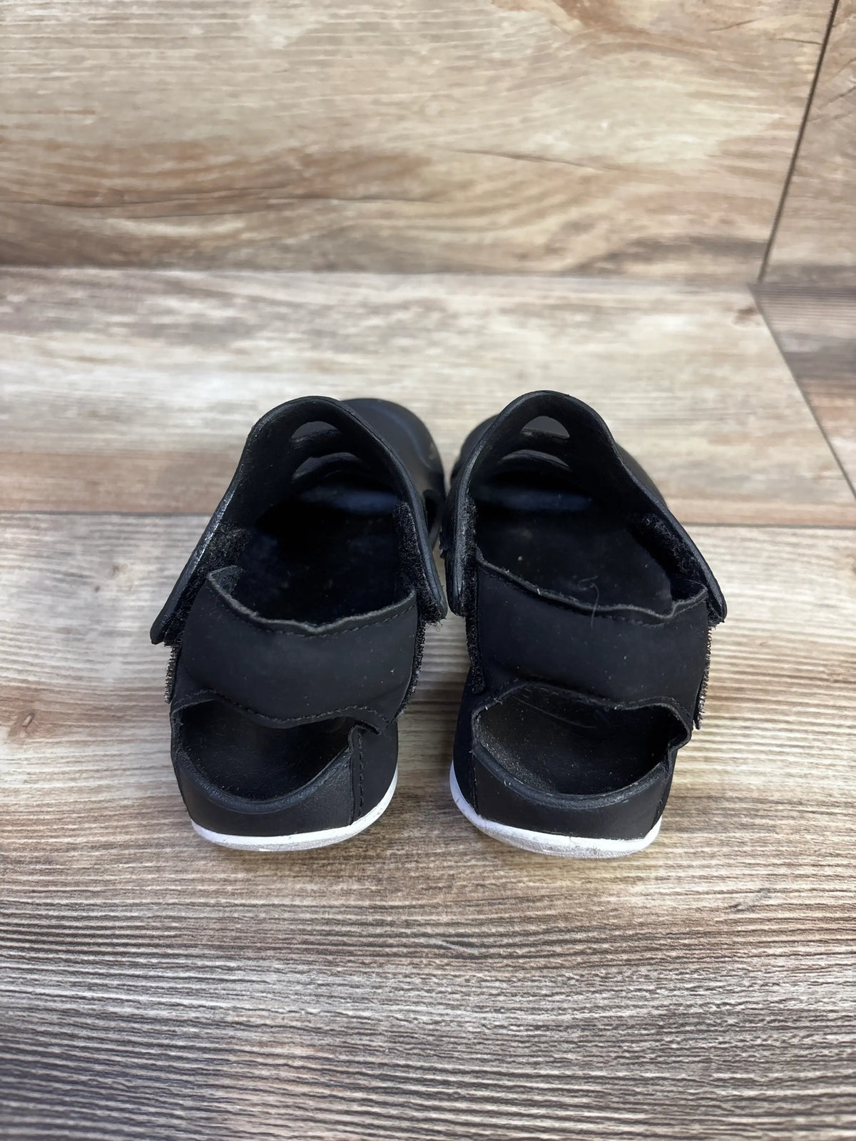 Rear view of Nike Sunray Protect 3 toddler sandals in black and white showcasing Velcro heel straps and white soles on wood background
