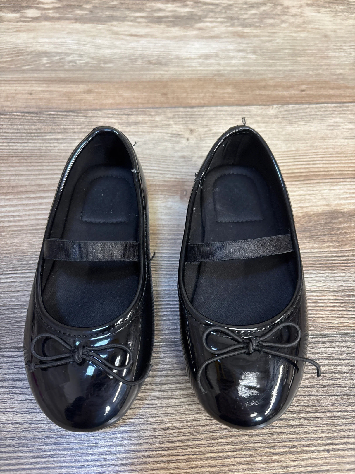 Top view of Jumping Beans Juniee Ballet Flat Black sz 8c showcasing patent black ballet flats with bow toe and elastic strap on wood plank floor