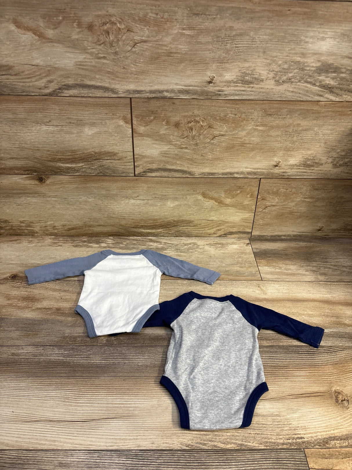 Flat lay of two newborn raglan bodysuits showcasing long raglan sleeves and blue and gray colorblocking on wood background