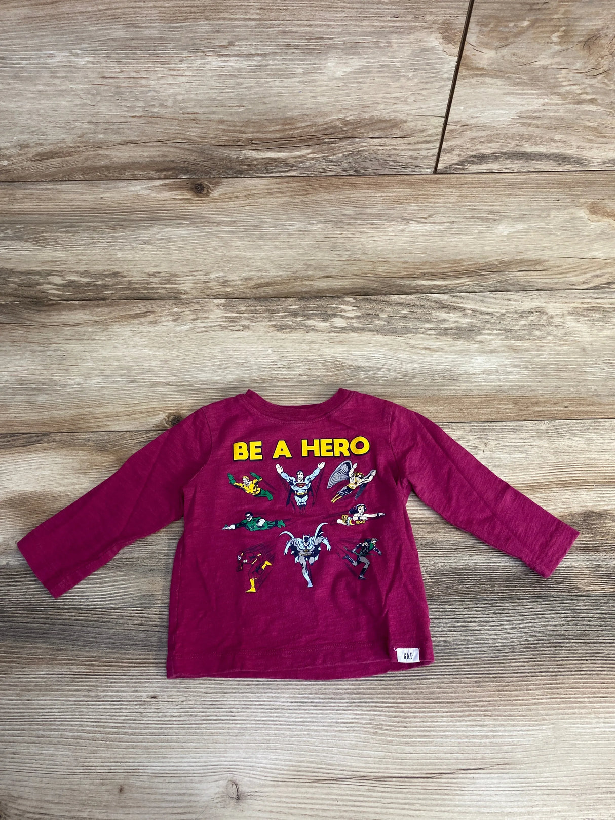 Flat-lay back view of Baby Gap infant long-sleeve T-shirt in cerise showing yellow BE A HERO and Justice League art on wood