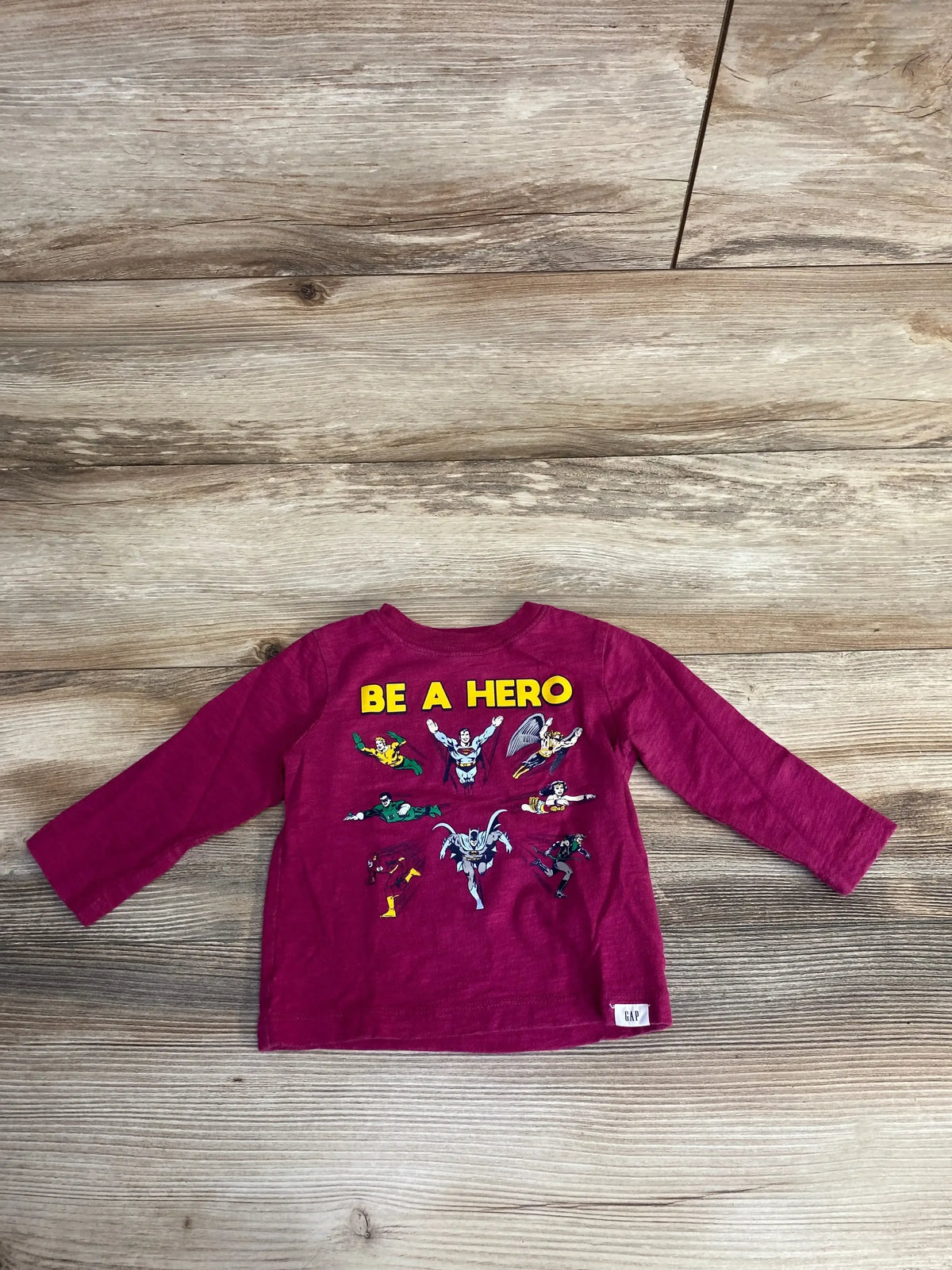 Flat-lay back view of Baby Gap infant long-sleeve T-shirt in cerise showing yellow BE A HERO and Justice League art on wood