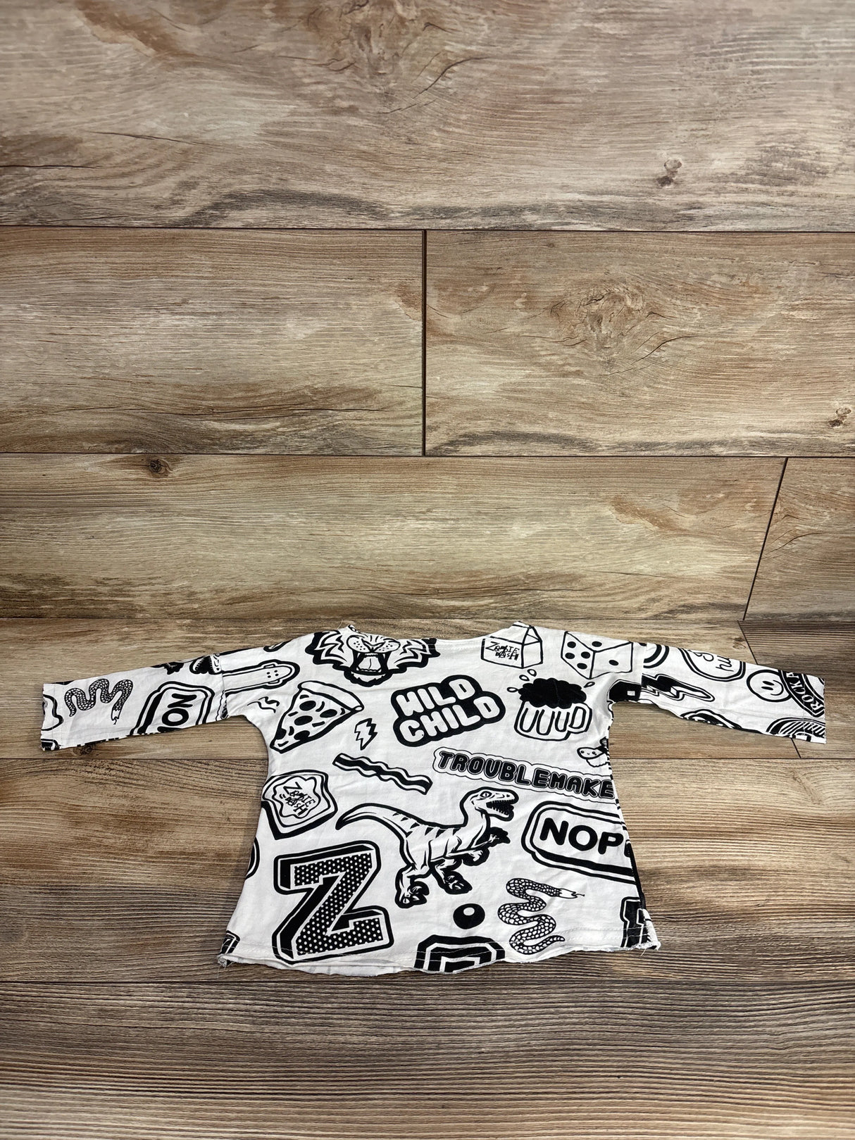 Kids long sleeve white shirt with black doodle graphics and text, laid on wood floor