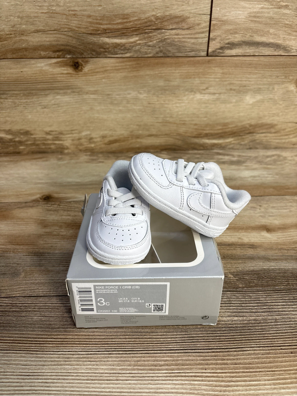Pair of white Nike Force 1 crib shoes on gray shoebox with wood background