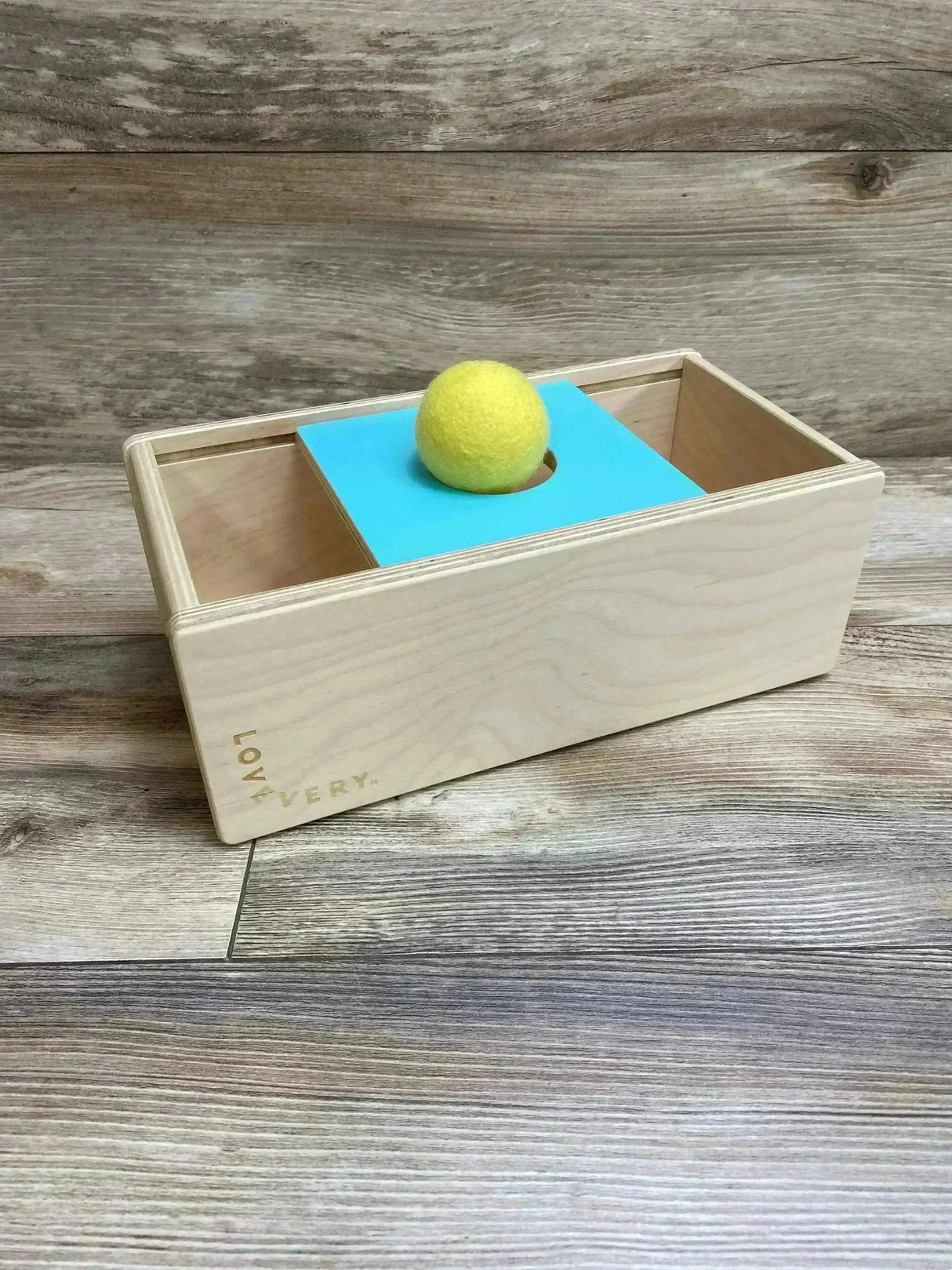 Three-quarter view of Lovevery Sliding Top Box showing turquoise sliding top with center hole and yellow felt ball on wood floor background