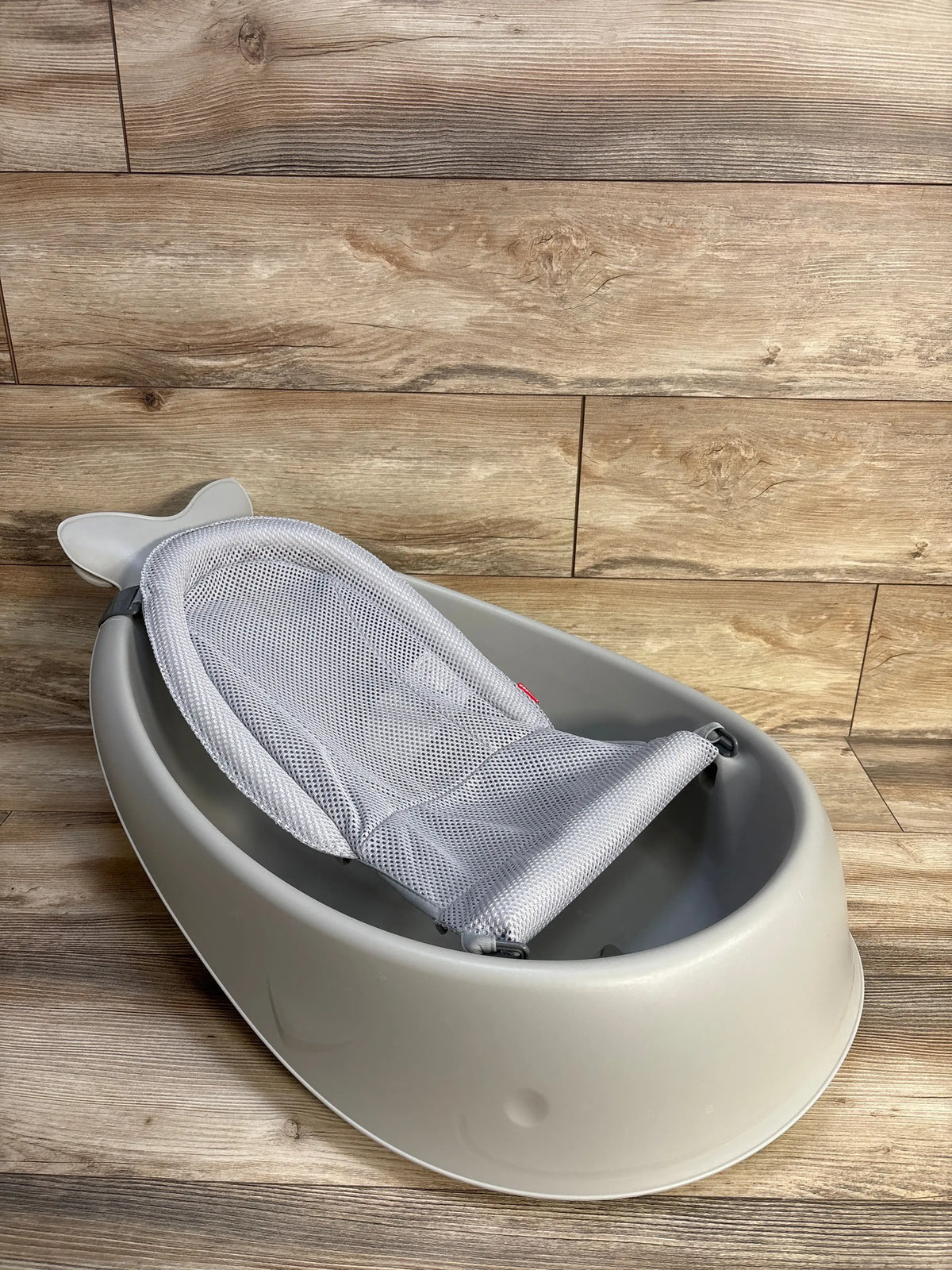 Angled view of Skip Hop Moby Smart Sling 3 in 1 baby bathtub showcasing its mesh Smart Sling with a wood-look floor background