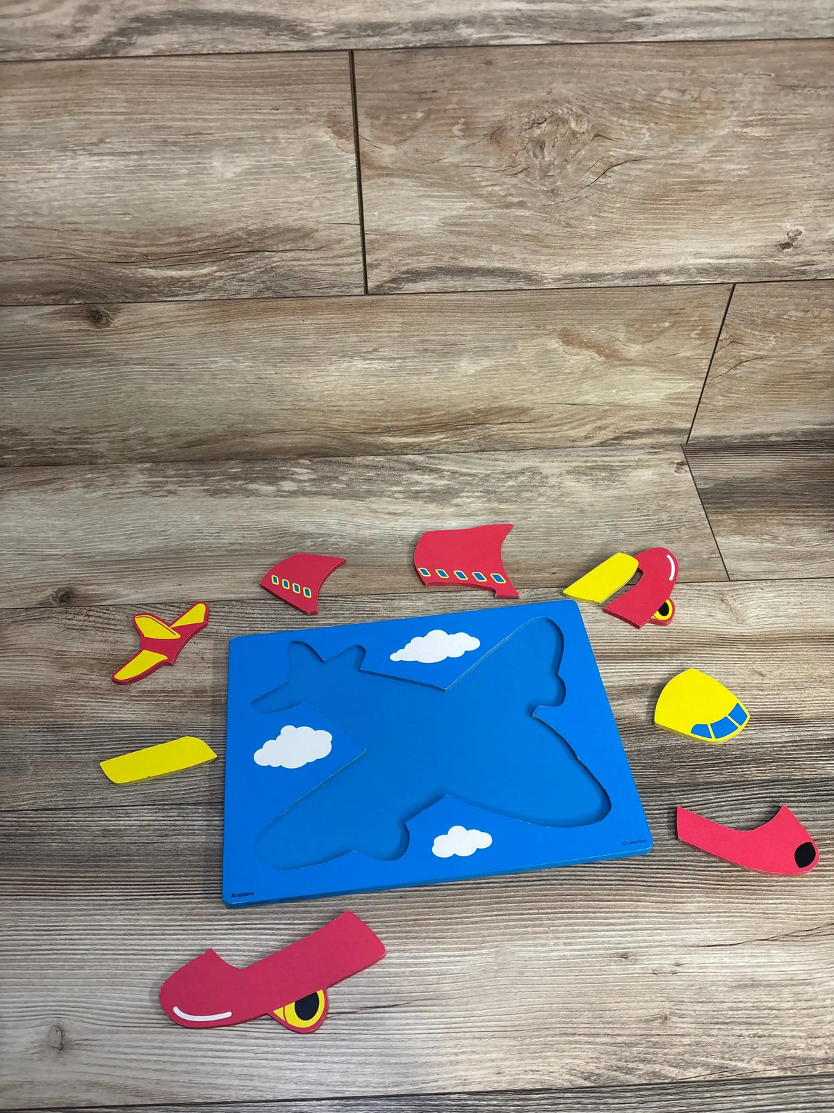 Top-down of Lakeshore 9-piece wooden airplane puzzle showing blue board with cloud illustrations and red and yellow plane pieces on wood floor