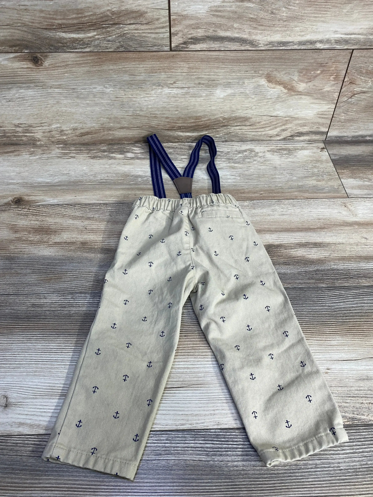 Flat lay of tan suspender pants with navy anchor print and navy striped elastic suspenders on wood background