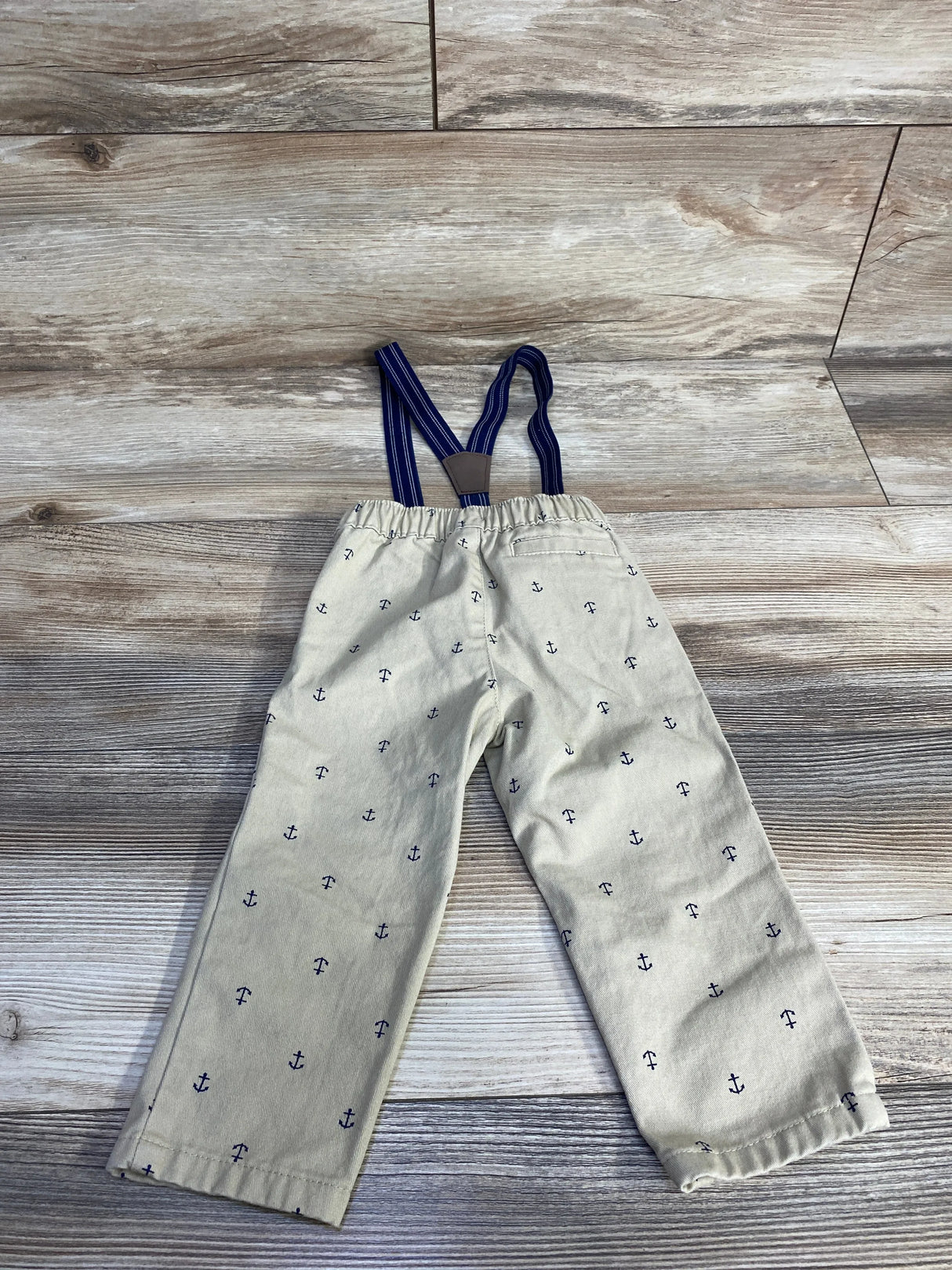Flat lay of tan suspender pants with navy anchor print and navy striped elastic suspenders on wood background