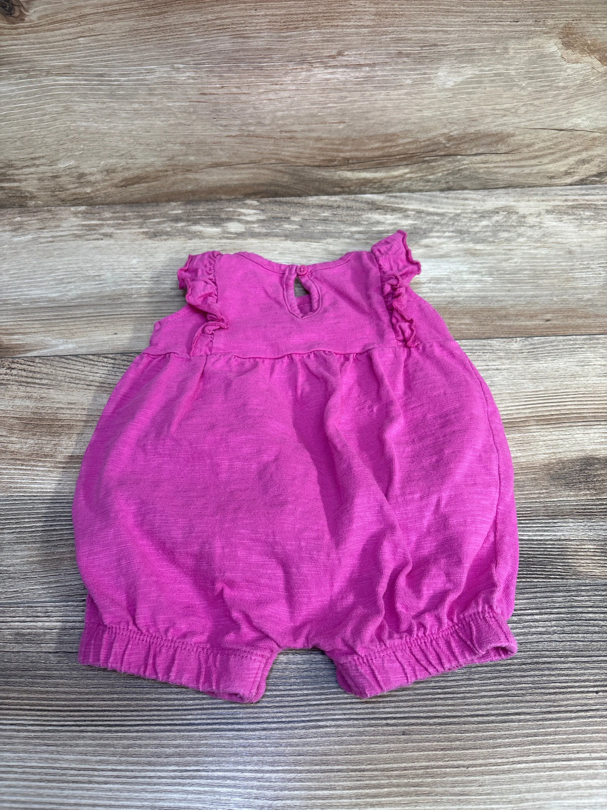Flat lay of pink baby ruffle shortie romper with ruffled sleeves, elastic leg openings and back keyhole on wood background