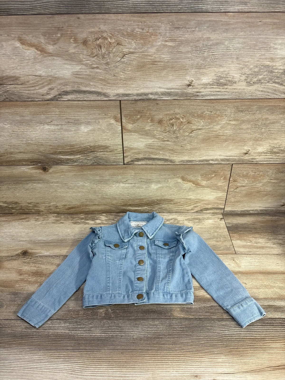 Front view of Btween light blue denim jacket, size 4T, showcasing ruffled shoulders and button closure on wood planks