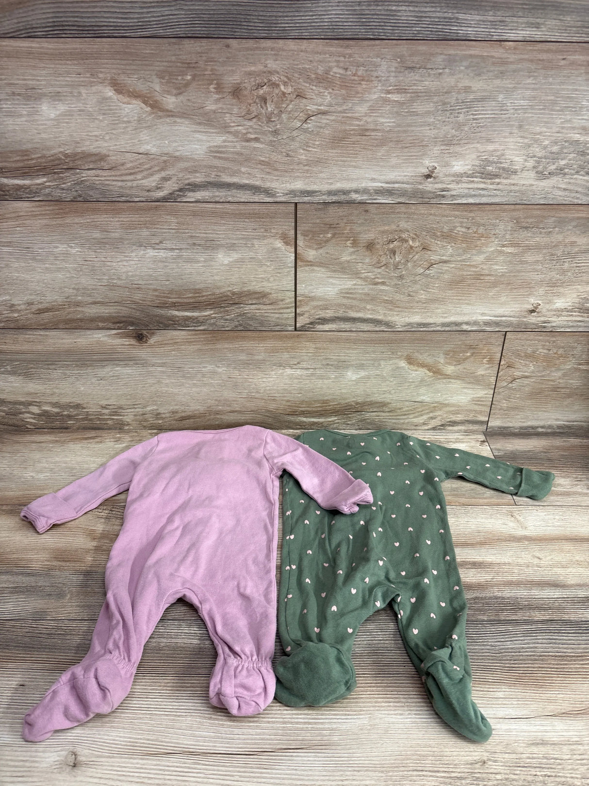 Flat lay of two newborn footed sleepers: mauve solid and green with tiny pink hearts, on light wood background