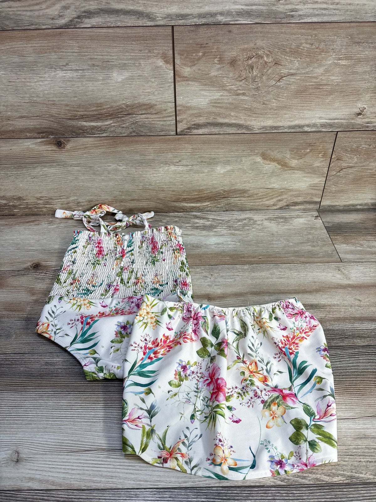 Flat lay of Jessica Simpson ivory two-piece smocked swimsuit and skirt set with floral print on a wood-look background