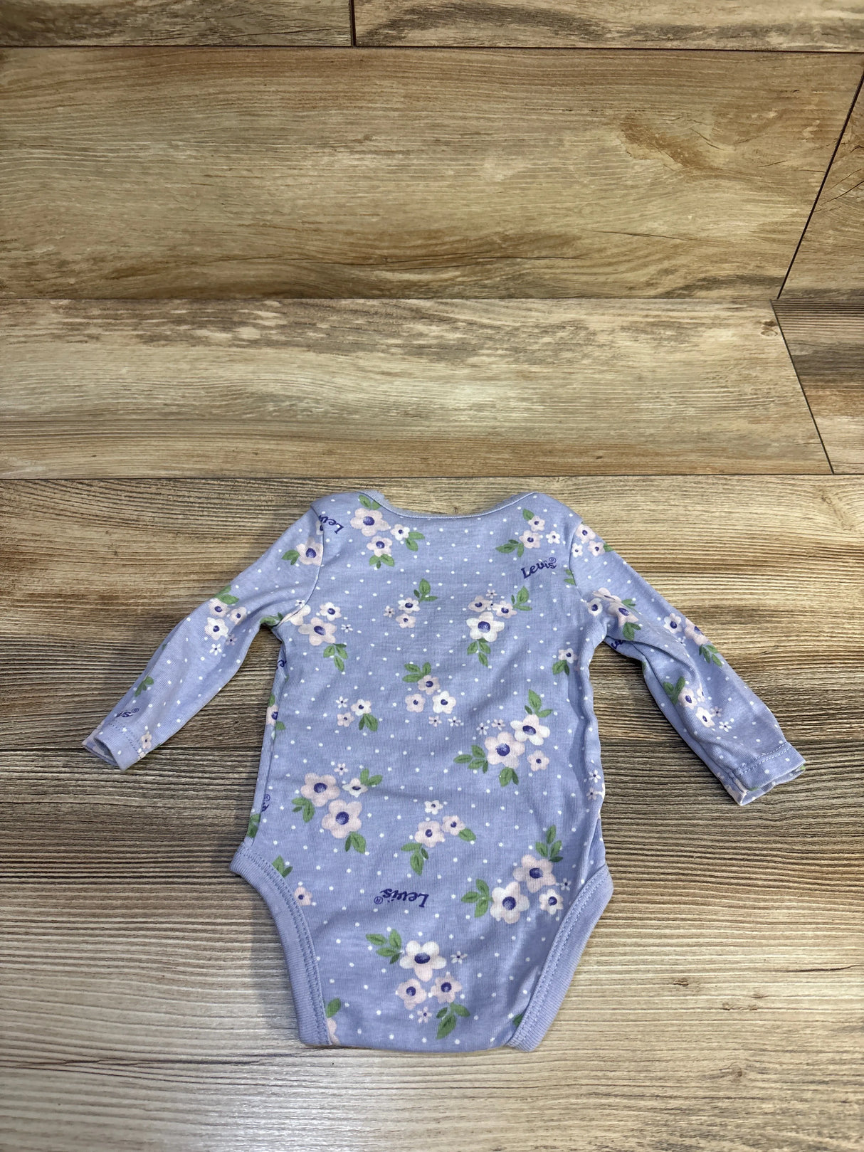 Flat lay of Levi’s lilac floral long-sleeve baby bodysuit with polka dots and snap crotch on wood floor, size 3 months