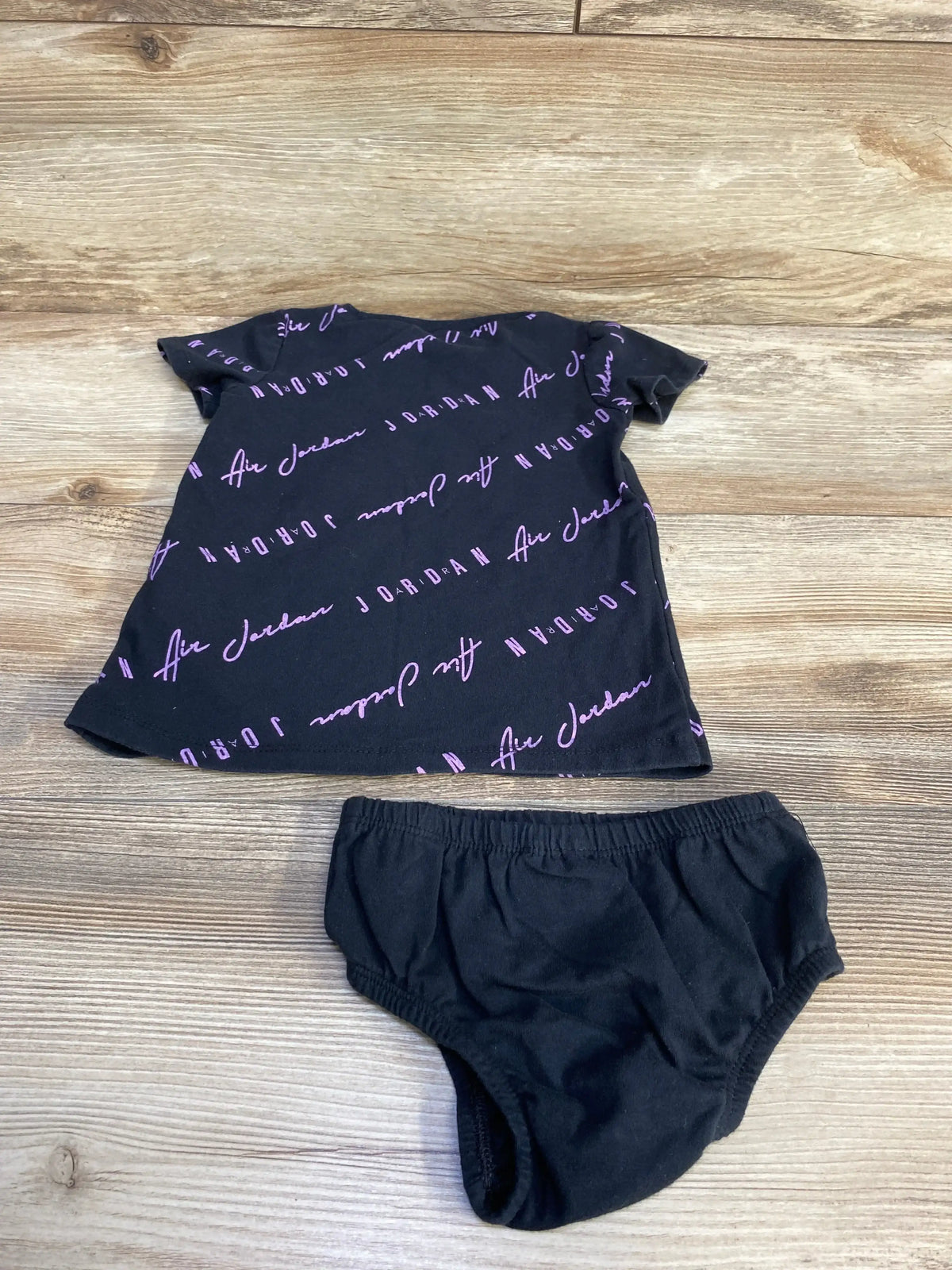 Front view of black Jordan 2-piece baby dress and bloomers showing purple diagonal Air Jordan script on wood background
