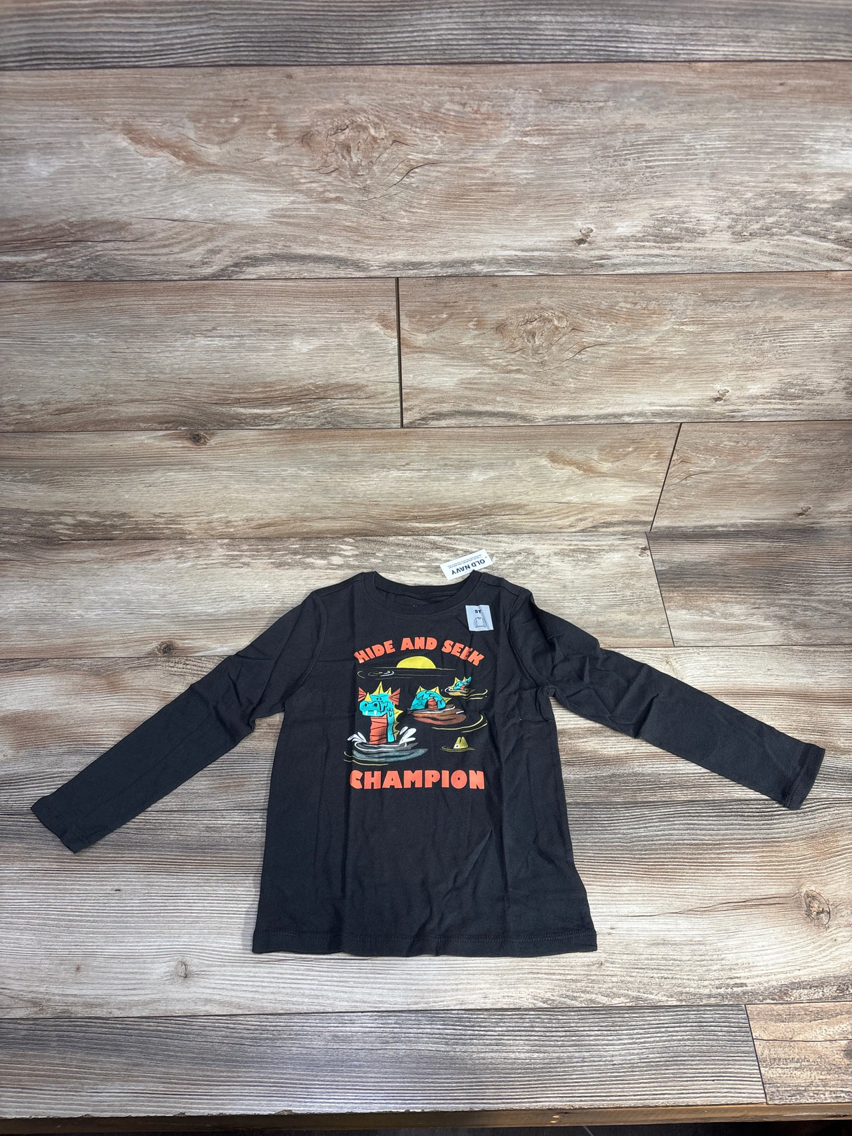 Black long sleeve kids shirt with cartoon cats and 'Hide and Seek Champion' text on wood floor