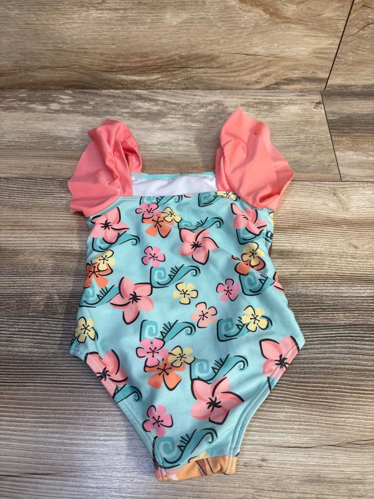Top view of 2T blue Moana one-piece swimsuit with coral ruffle sleeves and hibiscus floral print on wood background