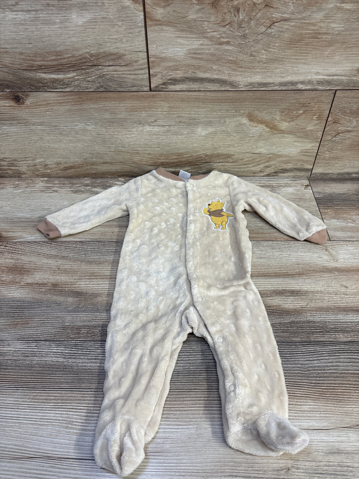 Front flat lay of 0-3M beige plush Winnie the Pooh footed sleeper with Pooh chest patch, front snaps and fold-over cuffs on wood background