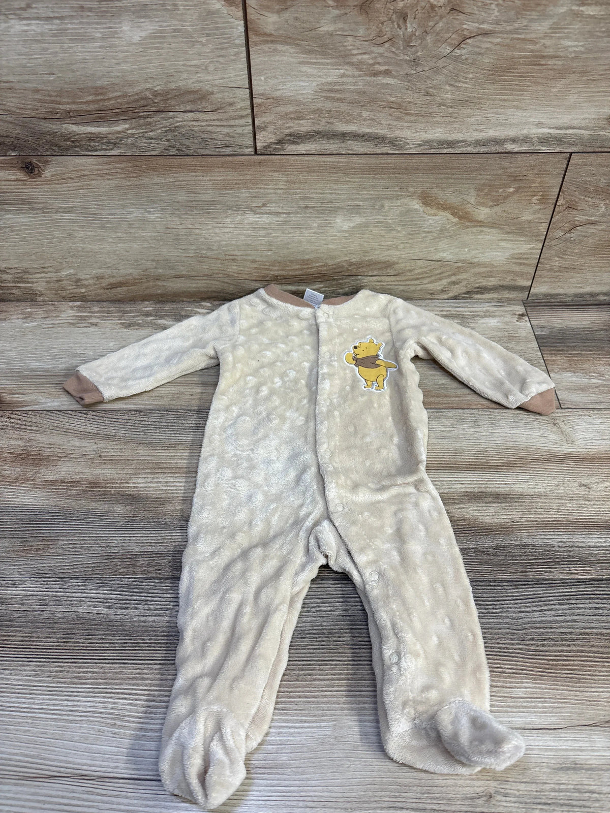 Front flat lay of 0-3M beige plush Winnie the Pooh footed sleeper with Pooh chest patch, front snaps and fold-over cuffs on wood background