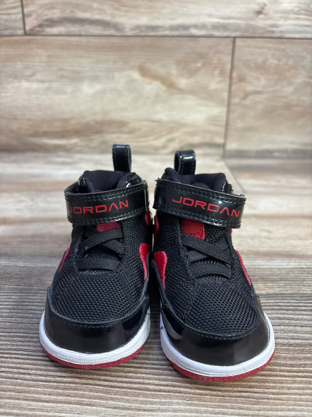 Front view of Jordan Flight TR 97 toddler sneakers, black with red accents, patent toe and velcro strap reading JORDAN