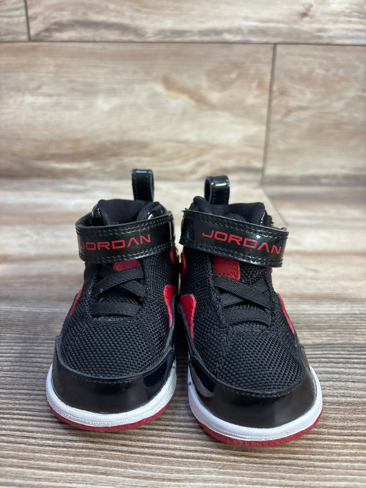 Front view of Jordan Flight TR 97 toddler sneakers, black with red accents, patent toe and velcro strap reading JORDAN